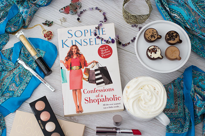 Confessions of a Shopaholic by Sophie Kinsella. Book around makeup, capuccino, jewellery, a scarf, chocolate cups.